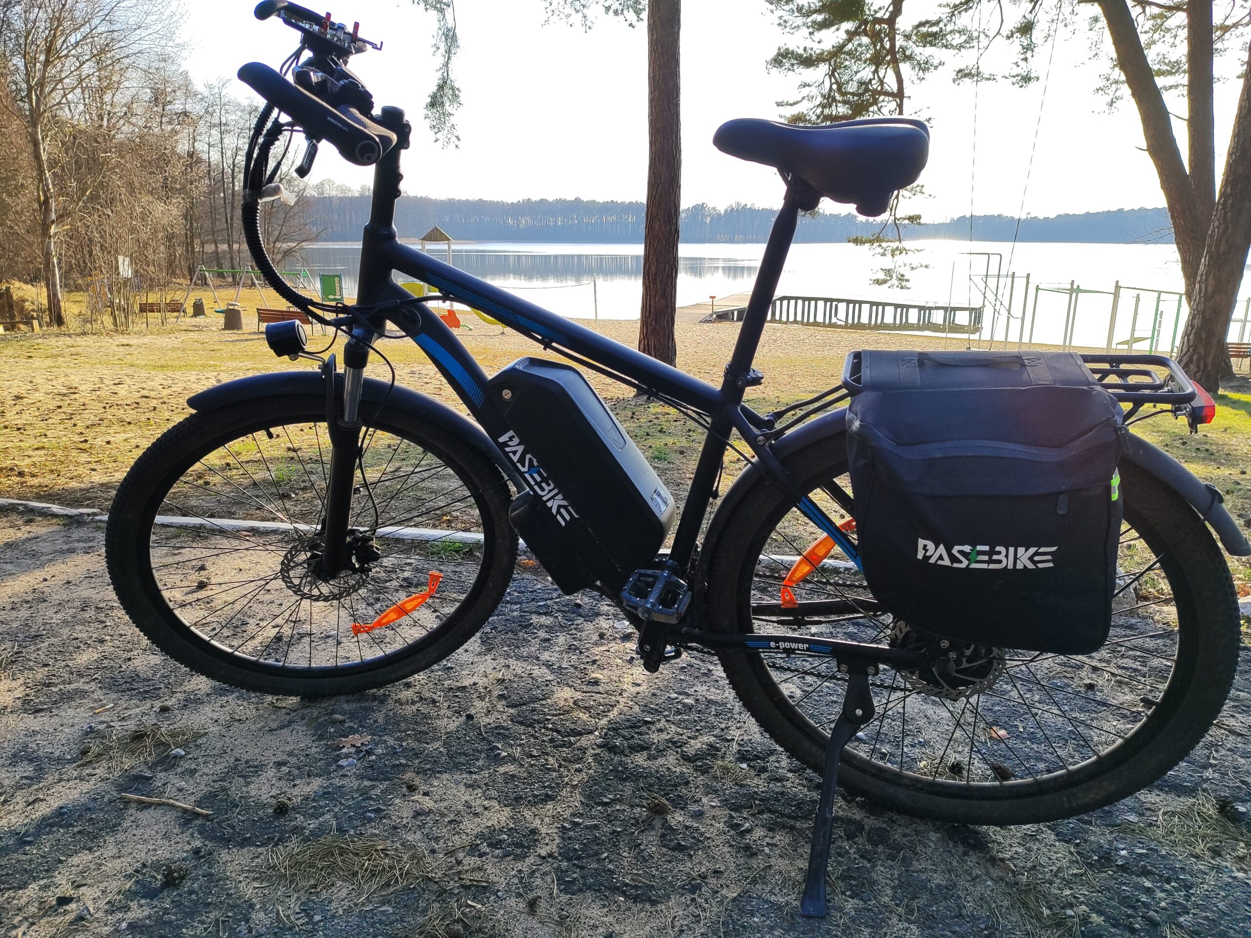 Rower elektryczny Pasebike C29 Pro z sakwami na bagażniku zaparkowany nad jeziorem w słoneczny dzień